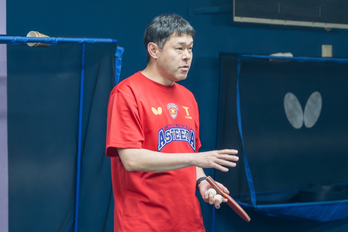 赤いアスティーダのシャツを着た田勢監督が、ラケットとボールを手に指導している様子